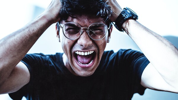 Man wearing glasses with hands on head, smiling widely with an energetic expression against a blue background.
