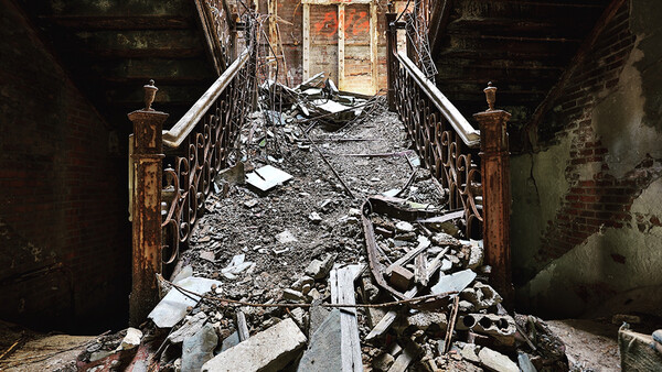 Abandoned stairwell filled with debris and rubble, with ornate metal railings and an open doorway at the far end.