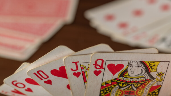 Playing cards fanned out showing hearts suit and a queen card, with additional cards blurred in the background on a dark wooden surface.