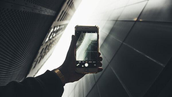 Hand holding smartphone capturing upward view of modern buildings against bright sky.