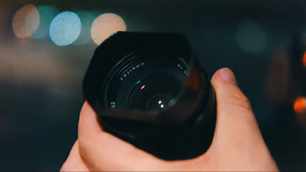 Hand holding a wide-angle camera lens with bokeh lights blurred in the background.