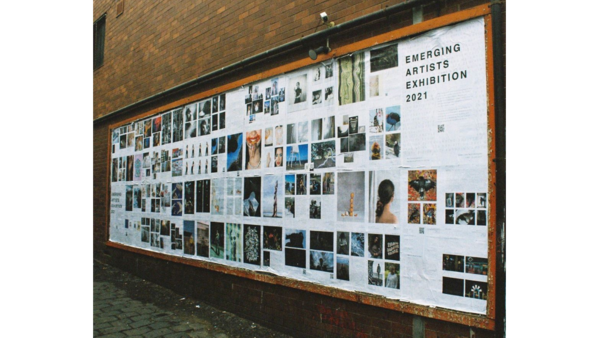Large outdoor gallery wall displaying numerous photographs and artworks arranged in a grid layout with an orange frame border.