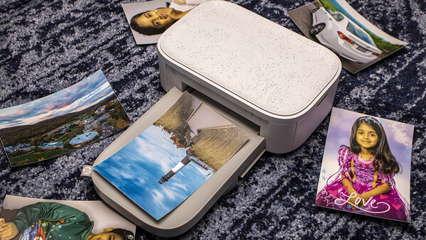 A white portable photo printer with printed photos and loose prints arranged on a dark textured surface.