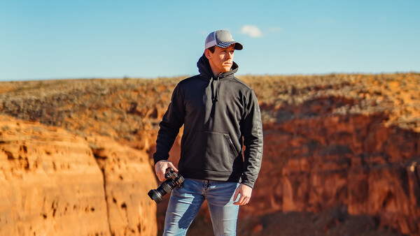 Man in cap and dark jacket holding camera stands at cliff edge overlooking layered canyon terrain.