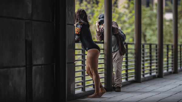 Woman standing on a wooden deck viewed through dark doorframe, with two people visible in background near railing.
