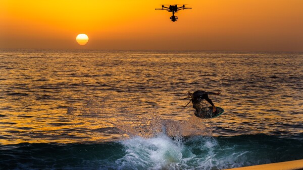 Surfer riding a wave at sunset with a drone flying overhead.
