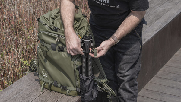 Person adjusting straps on an olive green camera backpack while standing on a wooden deck.