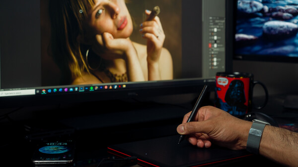 Photo editor's hand holding a stylus while working on a portrait image displayed on a monitor.