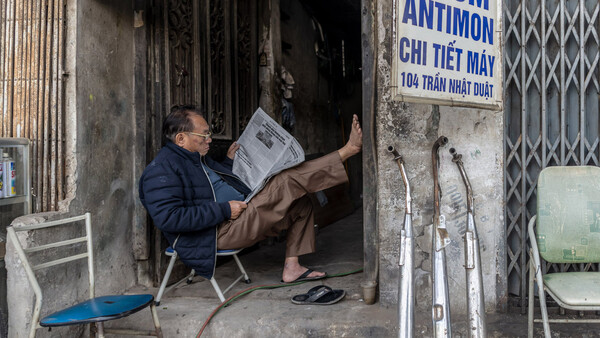 Man reading newspaper while sitting in doorway of weathered storefront with metal security gate.
