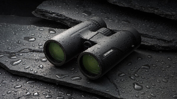 Black binoculars with yellow-green objective lenses on wet, dark textured surface with water droplets.