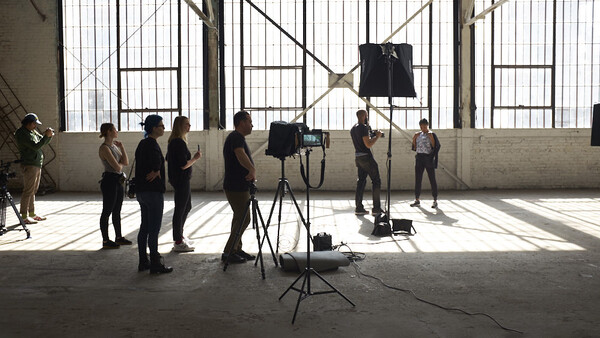 Film crew setting up camera and lighting equipment in an industrial studio space with large windows.