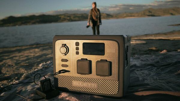 Portable power station with tan and black casing displayed on a beach, with a person standing in shallow water in the background.