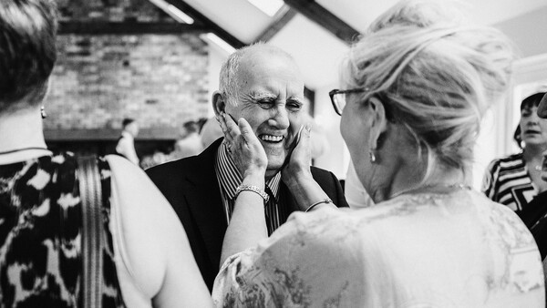 Elderly man with white hair smiling while touching his face during what appears to be a social gathering.