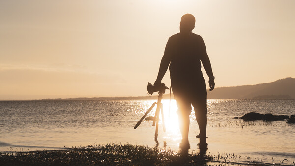 Silhouetted surfer carrying surfboard walking into ocean at golden hour.