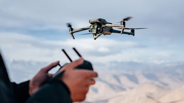 Drone operator holding remote controller with quadcopter flying overhead against cloudy sky and desert landscape.