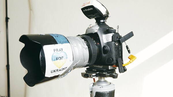 Professional video camera mounted on tripod with white tape reading "PRAY FOR UKRAINE" wrapped around lens barrel.