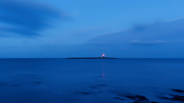 A solitary lighthouse beam cuts through deep blue twilight across a calm ocean horizon.