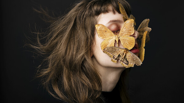 Profile of a woman with long dark hair and golden butterflies covering her eyes and mouth against a black background.