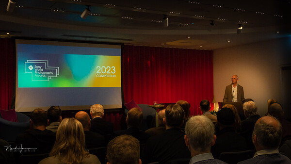 Conference speaker presenting to seated audience in auditorium with projection screen displaying 2023 event branding.