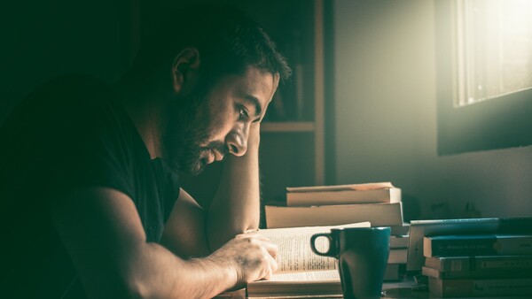 Man reading an open book at a desk with stacked books and a cup of coffee, lit by warm window light.