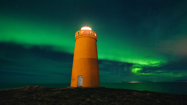 Bright orange lighthouse standing alone on dark terrain beneath vibrant green aurora borealis.