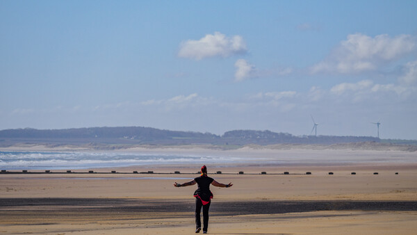 Person with arms outstretched standing alone on a wide, wet sandy beach with gentle waves and distant hills.
