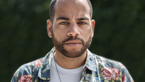 Man with beard wearing floral patterned shirt and white undershirt, photographed outdoors with blurred green background.