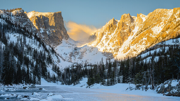 Frozen alpine lake surrounded by snow-covered peaks and coniferous forest at sunrise.