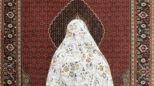 Woman in white patterned garment standing before ornate burgundy and gold traditional textile backdrop.