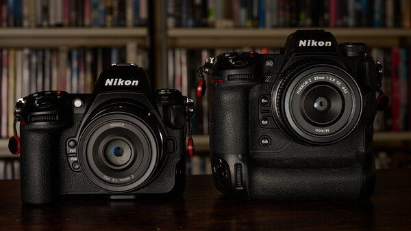 Two black Nikon DSLR cameras side by side, with the smaller model on the left and larger model on the right, displayed against a blurred bookshelf background.
