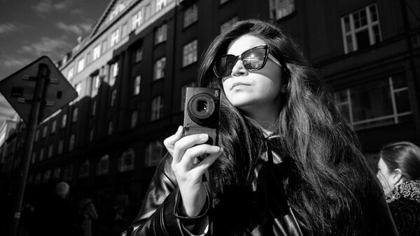 Woman in sunglasses holding a vintage camera on an urban street with period buildings behind her.