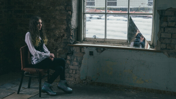 Person seated on a wooden chair in an industrial room, gazing toward a window overlooking a frozen landscape.