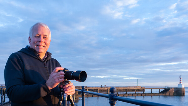 Photographer holding a telephoto lens camera on a tripod overlooking a harbor at golden hour.
