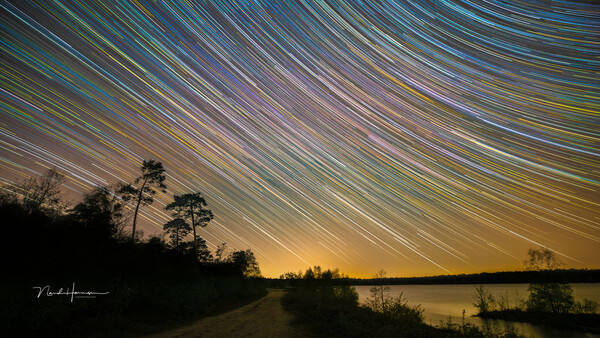 Star trail arcs sweep across the night sky above a dark lakeside landscape with silhouetted trees.