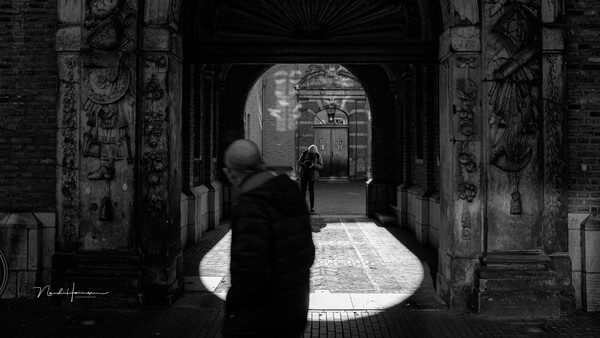 Person in dark clothing walking away through an ornate arched passageway toward distant figures in daylight.