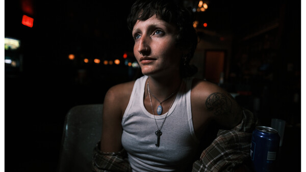 Person in white tank top and plaid shirt against dark background with warm ambient lights.