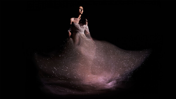 Woman in flowing, sparkle-embellished gown poses against dark background with dramatic lighting.