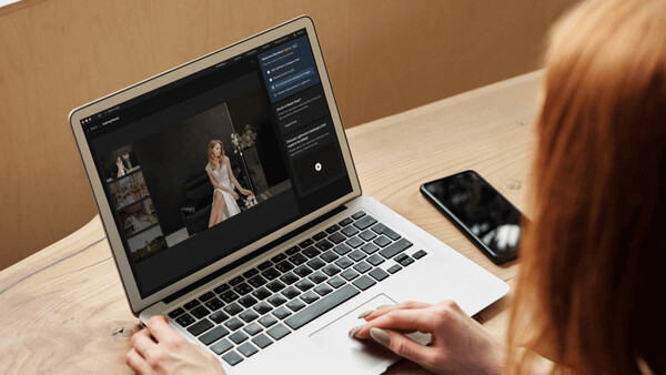 Photographer editing portrait images on laptop with smartphone nearby at wooden desk.