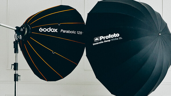 Two large parabolic umbrellas mounted on a stand: a Godox Parabolic 128 with gold interior and black exterior, and a Profoto umbrella with black exterior.