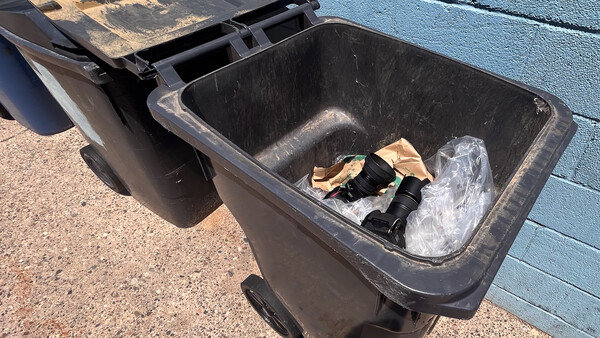 Two black wheeled trash bins against a blue wall, one partially open with crumpled plastic and cardboard inside.