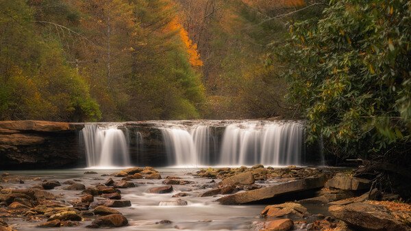 Cascading waterfall flowing over rocky ledges with autumn foliage in background.
