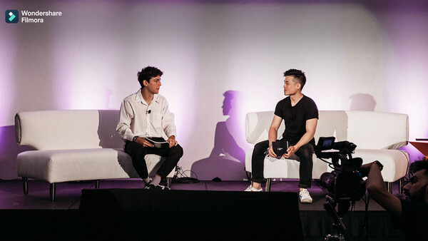 Two men sit on white couches facing each other during a staged interview or panel discussion with purple lighting.