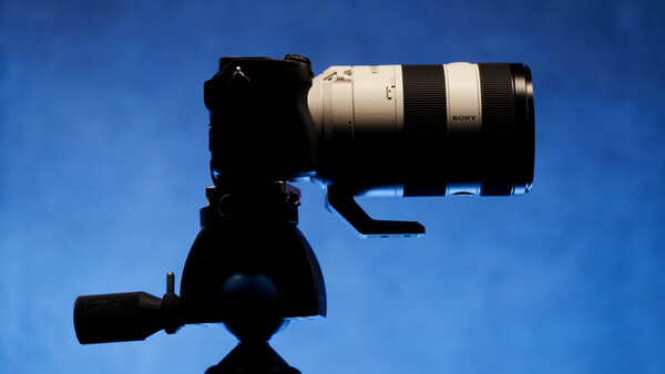 Silhouetted camera on tripod with telephoto lens mounted against a clear blue sky.