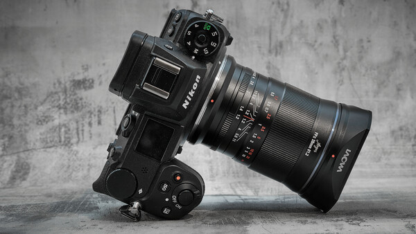 Mirrorless camera with telephoto lens mounted on motorized gimbal head against textured gray backdrop.