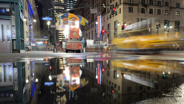 Wet city street at night with blurred yellow taxi and illuminated storefronts reflected in puddle.