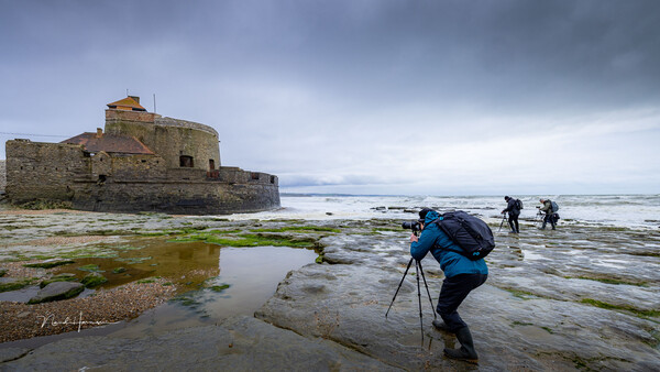 Photographer with tripod wading through tidal pools toward a stone fortress on a rocky coastline.