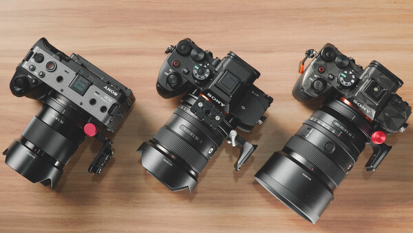 Three cinema cameras with large telephoto lenses arranged on a wooden surface, each equipped with top-mounted monitoring screens and red lens caps.