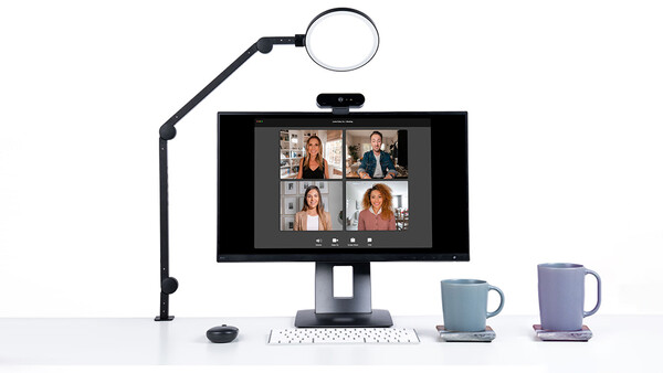 Video conferencing setup with monitor displaying four participants, ring light on articulating arm, keyboard, and two mugs.