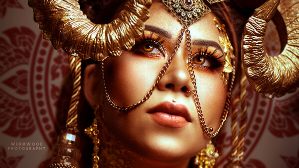 Woman wearing elaborate gold headdress and ornate jewelry with dramatic eye makeup against patterned backdrop.