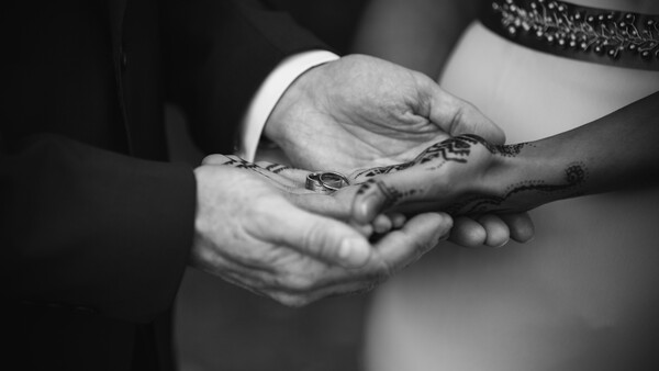 Two people's hands clasped together, one wearing a white sleeve and the other displaying henna designs on dark skin.